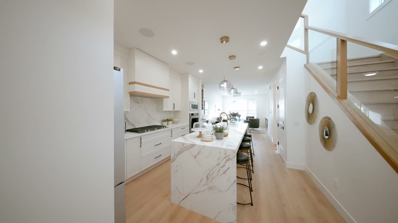 Listing film still: white kitchen with a waterfall island beside a glass staircase